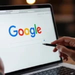 Egy felhasználó laptopon a Google keresőoldalát nézi, miközben egy tollal ír.