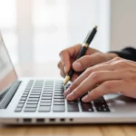 Egy kéz, amely egy laptop billentyűzetén gépel, miközben jegyzetel egy tollal, a tartalomfrissítés folyamatát szimbolizálva.