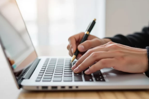 Egy kéz, amely egy laptop billentyűzetén gépel, miközben jegyzetel egy tollal, a tartalomfrissítés folyamatát szimbolizálva.