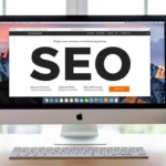 Egy asztalon elhelyezett számítógép képernyőjén a SEO kifejezés nagy betűkkel látható, háttérben színes minták és növények.
